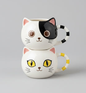 Two stacked ceramic cat-face mugs on a light gray background. The top mug has a black-and-white cat face with pink ears and a black-and-white striped handle.