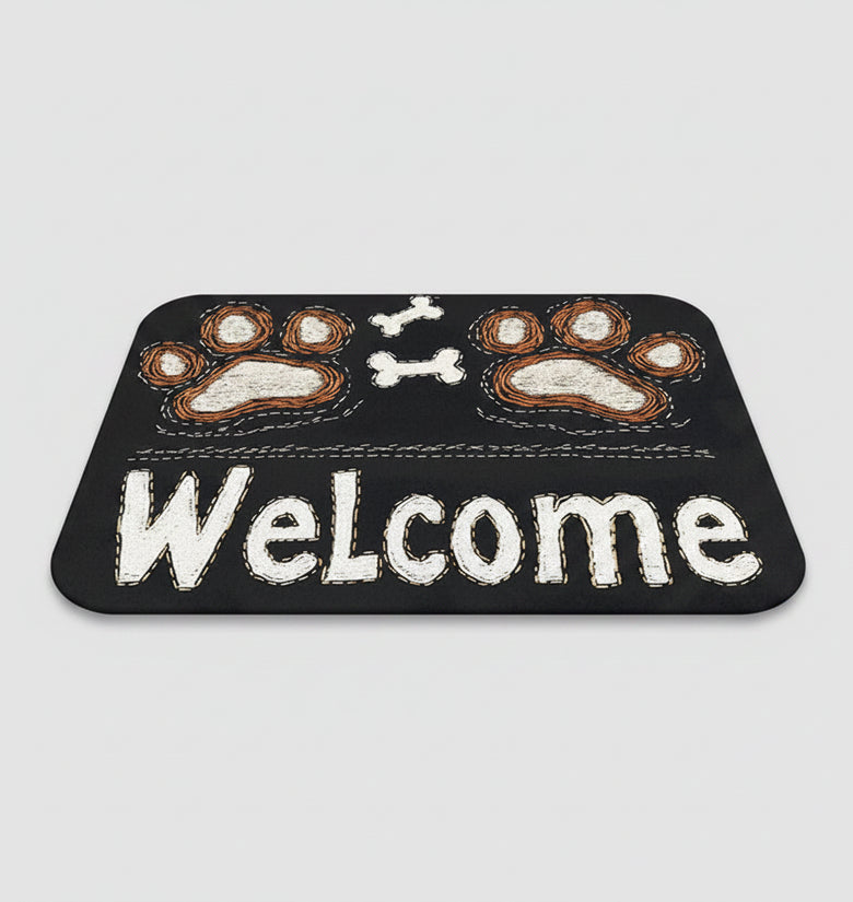 Black doormat with paw prints and 'Welcome' text on a wooden floor, next to a pair of slippers.