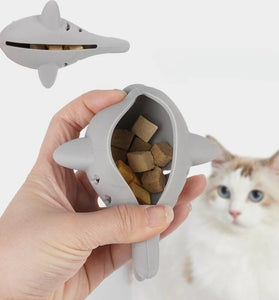 Gray shark-shaped pet toy with treats held by a hand, cat in the background