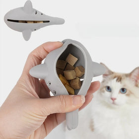 Gray shark-shaped pet toy with treats held by a hand, cat in the background